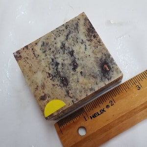 May include: A close-up of a square, light brown, gray, and black speckled stone with a yellow sticker on the corner. The stone is sitting on a wooden ruler marked with inches.