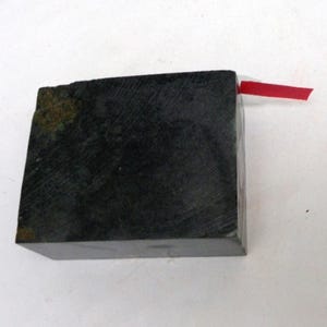 May include: A dark, rectangular block with a rough, textured surface. A small, bright red strip of material is attached to one edge. The block appears to be made of a dark stone or similar material.