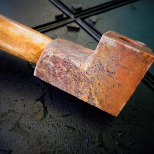 May include: A close-up of a pipe with a wooden stem and a square, stone bowl. The bowl has a mottled orange and brown pattern. The stem is a light tan color.