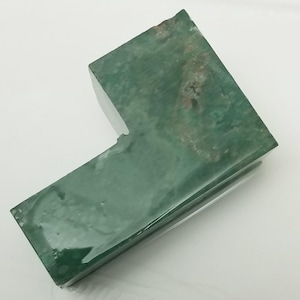May include: A green, L-shaped stone with a polished surface. The stone has a rough, textured area on one corner, with variations in color and tone. The object is set against a white background.
