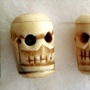 May include: Two small, white, carved skull beads. The beads have holes through the eyes and mouth.