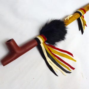 May include: A brown pipe with a decorative design. The pipe features a dark brown bowl and stem, connected to a wooden shaft. The shaft is adorned with black fur, colorful ribbons, and a bone-like accent.