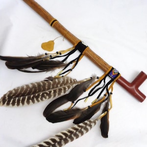 May include: A wooden ceremonial pipe with a brown finish, adorned with feathers, black and yellow ribbons, and beaded accents. The pipe features a T-shaped bowl and is decorated with various feathers.