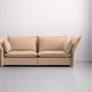 May include: A modern, two-cushion sofa in a neutral beige color. The sofa features large, square cushions and angled armrests, with additional smaller pillows. The room has white walls and a light-colored floor.