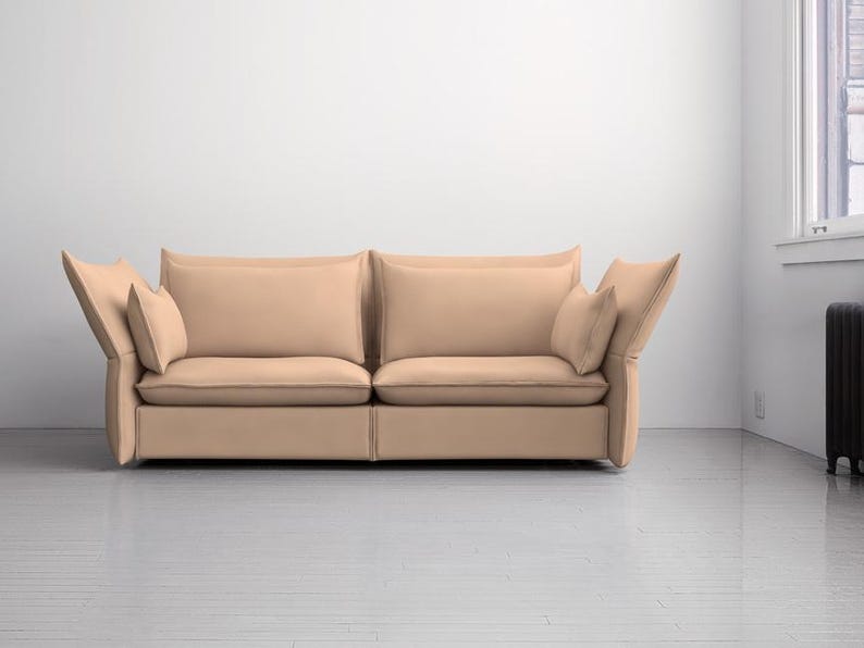 May include: A modern, two-cushion sofa in a neutral beige color. The sofa features large, square cushions and angled armrests, with additional smaller pillows. The room has white walls and a light-colored floor.