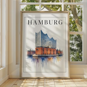 Op de afbeelding: Een ingelijste kunstdruk met een aquarelillustratie van de Elbphilharmonie in Hamburg, Duitsland. Het kunstwerk toont de rode bakstenen basis en de glazen bovenbouw met het woord HAMBURG in zwarte hoofdletters gedrukt.