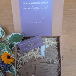 Könnte beinhalten: Eine Geschenkschachtel aus Karton mit einer lila Karte mit dem Text Manchmal fehlen einfach die richtigen Worte, einem Perlenarmband auf einer Karte, einem runden weißen Stein mit deutschem Text und mehreren kleinen braunen Karten.