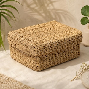 Puede incluir: Una caja de almacenamiento rectangular con tapa, fabricada con fibras naturales trenzadas de color beige claro. La caja presenta una textura trenzada consistente en todo el exterior, incluyendo la superficie superior y los lados verticales.