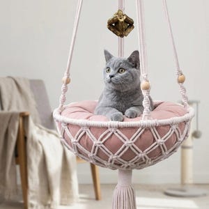 May include: A gray cat resting in a hanging macrame pet bed. The bed features a round, dusty pink cushion held by cream-colored knotted cotton cords with wooden beads. A small, textured wooden toy hangs from the top center of the suspension ropes.