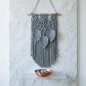 May include: A grey macrame wall hanging featuring three leaf-shaped elements suspended from a horizontal wooden dowel. The design incorporates intricate knotted patterns and long, vertical fringe strands that extend downward from the leaves.