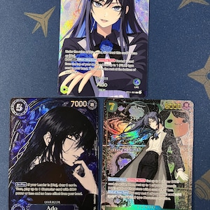 Peut inclure: Trois cartes à collectionner One Piece représentant le personnage Ado. La carte du haut montre Ado avec des cheveux bleus et une rose bleue. La carte en bas à gauche montre Ado aux longs cheveux noirs et roses bleues. La carte en bas à droite montre Ado en manteau.