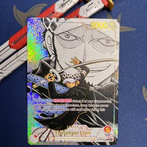 May include: A holographic One Piece trading card featuring Trafalgar Law, with a power level of 5000 and 5 life points. The card displays text detailing game mechanics and is positioned in front of two red, white, and gold toy swords.