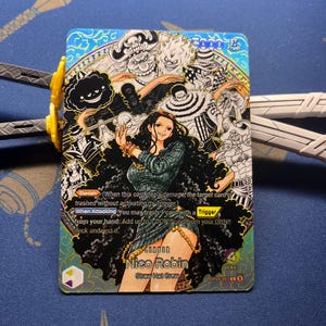 May include: A One Piece trading card featuring Nico Robin as the leader, with a power of 5000 and 4 life. The card displays a central illustration of Nico Robin surrounded by various characters in a black and white style, with blue and gold accents.