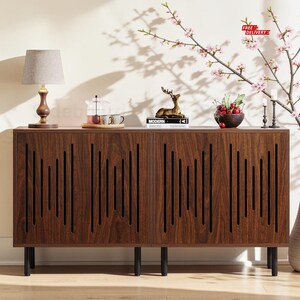 May include: A rectangular wooden sideboard featuring two cabinet doors with vertical slat cutouts, supported by four black cylindrical legs. The top surface holds a table lamp with a grey shade, a glass teapot, a deer figurine, and a bowl of cherries.