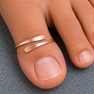 Könnte beinhalten: Ein offener Zehenring in Roségold mit einem glatten, minimalistischen Banddesign und abgerundeten Enden. Der Metallring wird am großen Zeh getragen und weist eine polierte Oberfläche auf, die das Licht auf seiner geschwungenen Form reflektiert.