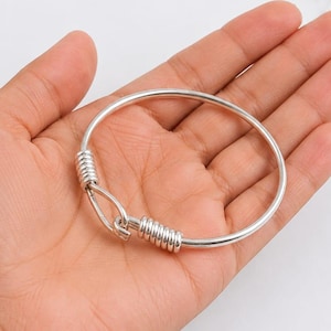 May include: A silver-toned bangle bracelet featuring a hook-and-eye style closure mechanism. The circular band is constructed from a smooth, polished metal wire, with coiled wire detailing accentuating the hook clasp at the center point.