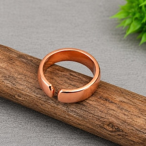 May include: A polished copper-colored ring with a smooth, rounded surface and an open-ended design. The band features a consistent width throughout its circumference, showcasing a metallic finish that reflects light across its curved exterior.
