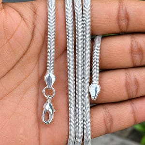 Op de afbeelding: Een zilverkleurige slangenketting met een glad, flexibel en strak geweven visgraatpatroon. De ketting is voorzien van een gepolijst druppelvormig eindstuk en een veilige karabijnsluiting om de ketting vast te maken.