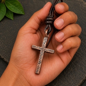 May include: A copper-toned metal cross pendant with a heavily hammered, textured surface. The cross is suspended from a dark brown leather cord that is knotted at the top, creating a rustic appearance with visible metallic oxidation and dark patina.