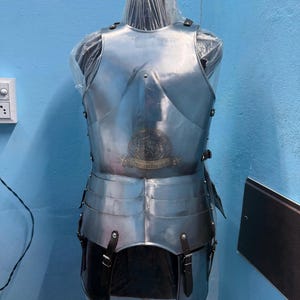 May include: A polished steel breastplate armour set featuring a central chest plate, layered tassets, and dark brown leather straps with metal buckles. A circular logo with the text Crescent Barakah is visible on the front of the chest piece.