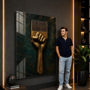 May include: A large rectangular wall art piece featuring a textured, three-dimensional gold-colored fist holding an afro pick comb. The artwork is set against a dark, mottled green and black background with a glossy finish, mounted on a dark wall.