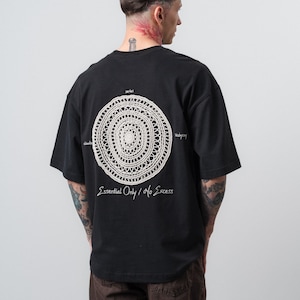 May include: A black short-sleeve t-shirt featuring a large, circular, cream-colored crochet applique on the back. The design includes small white text around the circle and the phrase "Essential Only / No Excess" printed in white below it.
