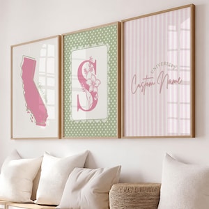 May include: Three framed wall art prints in light wood frames. The left print shows a pink California map outline with the text Your State. The center print features a large pink letter S with floral accents on a green polka dot background. The right print has pink vertical stripes with the text University Custom Name.
