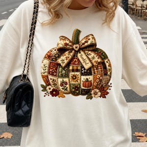 Peut inclure: Un sweat-shirt à col rond couleur crème arborant un grand motif de citrouille style patchwork. La citrouille est décorée de motifs floraux, d'un nœud à carreaux, d'un petit bouton en forme de cœur et d'une étiquette avec le mot thankful.