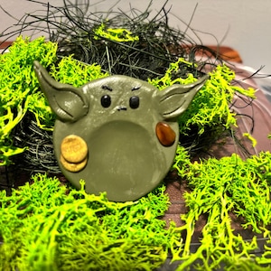 May include: A circular olive green clay sculpture featuring two pointed ears, small black eyes, and a small mouth. The sculpture has a smooth surface with a light brown circular patch on the left side and a dark brown circular patch on the right side.