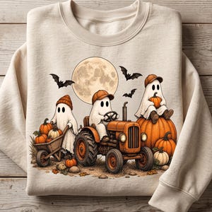 Può includere: Una felpa girocollo beige con un'illustrazione grafica di tre fantasmi bianchi con cappelli arancioni. Un fantasma guida un trattore arancione, un altro spinge una carriola di legno piena di zucche e il terzo è seduto sopra una grande zucca.