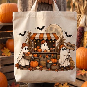 Può includere: Una borsa in tela color crema con un'illustrazione grafica di tre fantasmi con berretti arancioni presso una bancarella di zucche. Il disegno include zucche, steli di mais, pipistrelli, una luna piena e un cartello con la scritta PUMPKINS.