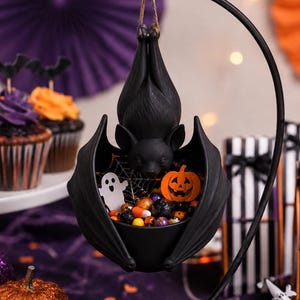 Könnte beinhalten: Eine schwarze, fledermausförmige Schale, die an einem geschwungenen Metallständer hängt. Die Schale zeigt oben eine modellierte Fledermausfigur und enthält kleine Halloween-Dekorationen, darunter ein weißes Gespenst, einen Kürbis und bunte Bonbons.
