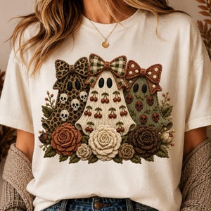 Può includere: Una t-shirt color crema con una grafica di tre figure a forma di fantasma adornate con fiocchi. I fantasmi sono decorati con motivi di teschi, ciliegie e pois, circondati da un gruppo di fiori e foglie in stile uncinetto.
