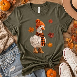 Può includere: Una t-shirt verde oliva con la grafica di un'oca bianca lavorata a maglia che indossa un berretto di lana arancione, una sciarpa a quadri neri e arancioni e stivaletti di lana arancioni abbinati, circondata da foglie autunnali sparse.