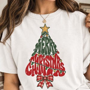 May include: A white short-sleeve t-shirt featuring a graphic of a Christmas tree formed by the text Merry Christmas in green and red. The tree is topped with a gold star, wrapped in string lights, and finished with a red and white plaid bow at the base.