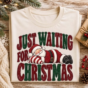May include: A cream-colored t-shirt featuring a graphic of Santa Claus sleeping on a striped pillow. The text "JUST WAITING FOR CHRISTMAS" is printed in large, bold, red and green letters surrounding the illustration of the resting Santa.