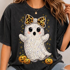 Può includere: Una t-shirt a maniche corte grigio antracite con una grafica centrale raffigurante un fantasma bianco con grandi occhi neri e guance rosa. Il fantasma indossa un fiocco leopardato ed è circondato da stelle dorate e tre piccole zucche arancioni.