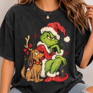 May include: A charcoal gray t-shirt featuring a graphic print of a green character wearing a red Santa hat and coat, sitting next to a brown dog with a red collar and a single antler attached to its head, surrounded by small red and green stars.