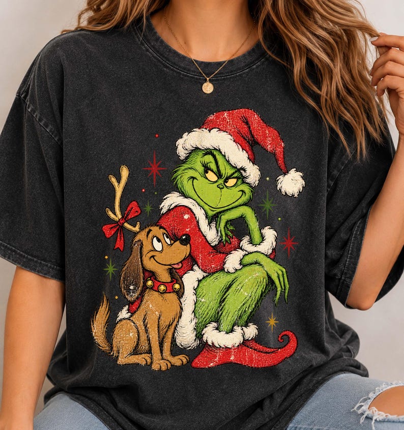May include: A charcoal gray t-shirt featuring a graphic print of a green character wearing a red Santa hat and coat, sitting next to a brown dog with a red collar and a single antler attached to its head, surrounded by small red and green stars.