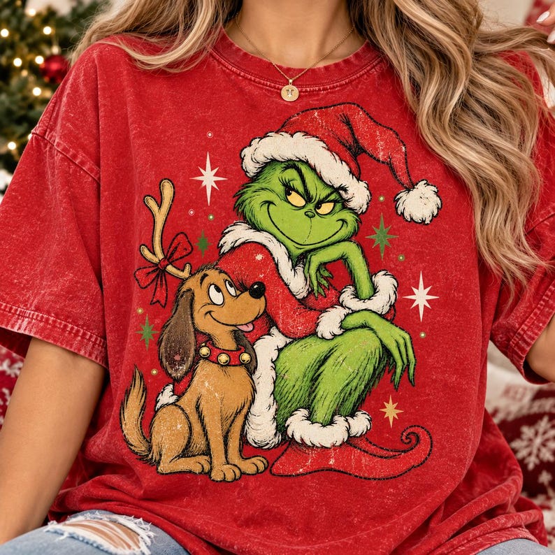 May include: A red mineral-washed t-shirt featuring a graphic print of a green creature wearing a Santa hat and coat, sitting next to a brown dog with reindeer antlers. The design includes small white and green star accents on the fabric.