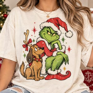 May include: A cream-colored t-shirt featuring a graphic print of a green character wearing a red Santa hat and coat, sitting next to a brown dog with reindeer antlers and a red collar with bells, surrounded by red and green star-shaped accents.
