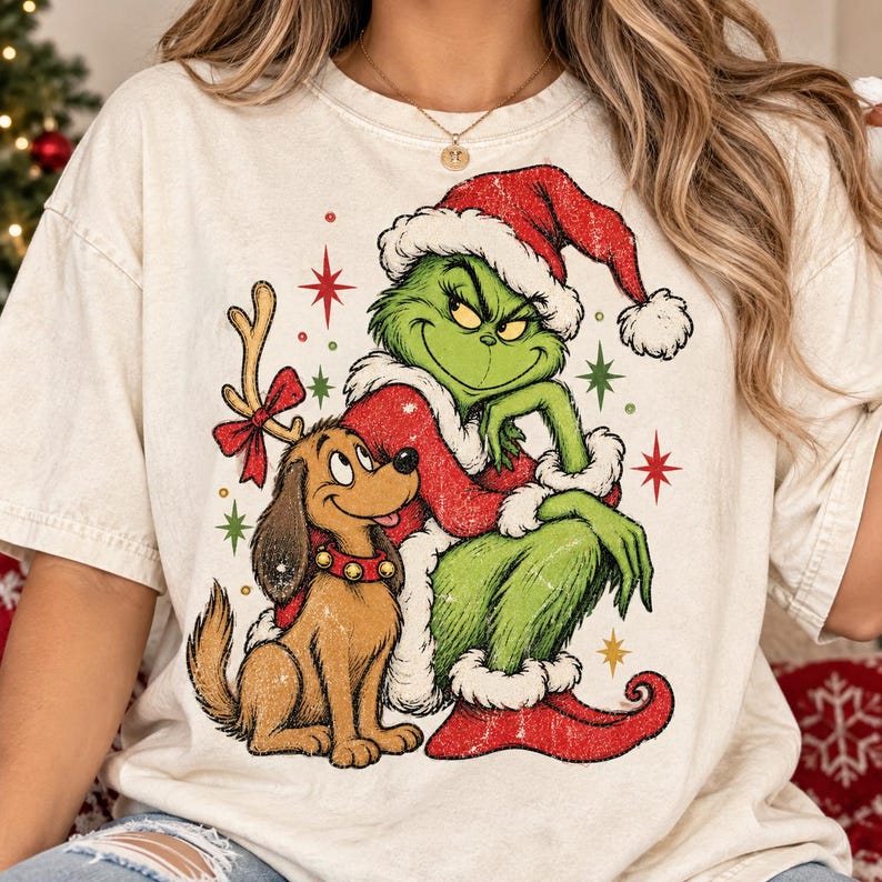 May include: A cream-colored t-shirt featuring a graphic print of a green character wearing a red Santa hat and coat, sitting next to a brown dog with reindeer antlers and a red collar with bells, surrounded by red and green star-shaped accents.