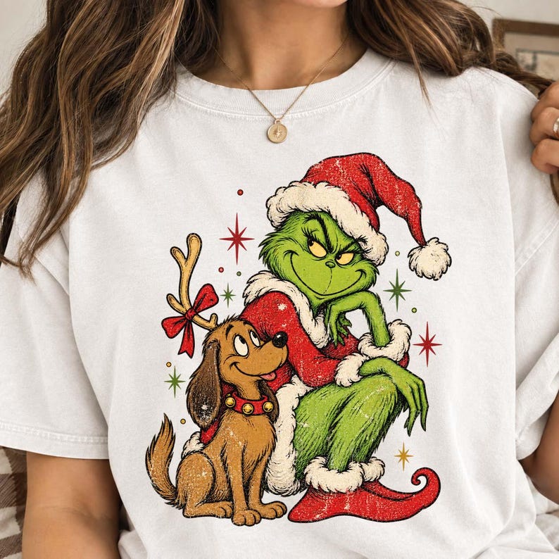 May include: A white crewneck t-shirt featuring a graphic print of a green character wearing a red Santa hat and coat, sitting next to a brown dog with reindeer antlers and a red bow. The design includes small red, green, and yellow star accents.