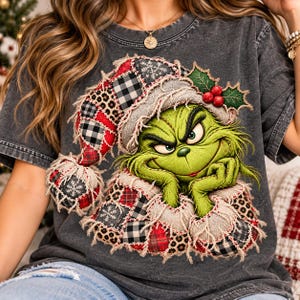 May include: A charcoal gray t-shirt featuring a graphic of a green creature wearing a patchwork Santa hat. The hat and base are decorated with red, black, and white plaid, leopard print, and snowflake patterns, accented with holly berries.