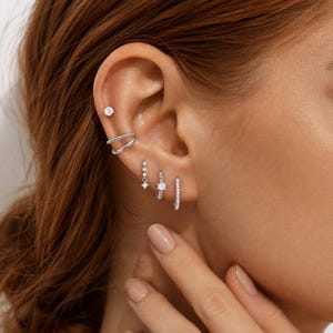 Könnte beinhalten: Ein Set aus silberfarbenen Ohrringen und Ohrklemmen mit klaren Steinen. Die Kollektion umfasst einen kleinen Stecker, eine doppelte Ohrklemme, einen hängenden Ohrring mit sternförmigem Stein, einen kleinen Ring mit Stein und einen Pavé-Ring.