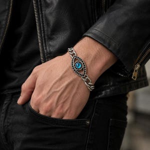 Könnte beinhalten: Ein silberfarbenes Gliederarmband mit einem zentralen, augenförmigen Anhänger. Der Anhänger hat ein dunkles, oxidiertes Metallfinish mit einem leuchtend blauen, runden Stein in der Mitte, der an eine Iris erinnert. Das Armband hat einen Metallverschluss.