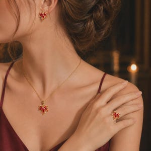 Könnte beinhalten: Ein Goldschmuckset bestehend aus einer Halskette, einem Paar Ohrringen und einem Ring, die jeweils ein Ahornblatt-Design mit leuchtend roten Steinen aufweisen. Die Halskette hat eine zarte Goldkette, passend zu Ring und Ohrringen.