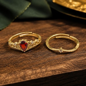May include: A set of two gold-toned adjustable rings. One ring features a crown-like design with a central pear-shaped red gemstone and small clear stones. The second ring is a thin band with a single small clear stone at the center.