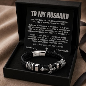 May include: A black braided leather bracelet with silver-toned metal accents, a cross charm, and a clasp engraved with the name Bryce. It is presented in a black gift box with a card featuring the text TO MY HUSBAND and a sentimental message.