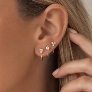 Könnte beinhalten: Drei goldene Creolen an einem Ohrläppchen. Der erste Ohrring hat einen birnenförmigen klaren Stein, der zweite einen runden klaren Stein und der dritte eine Anordnung aus drei kleinen runden klaren Steinen in einer dreieckigen Form.